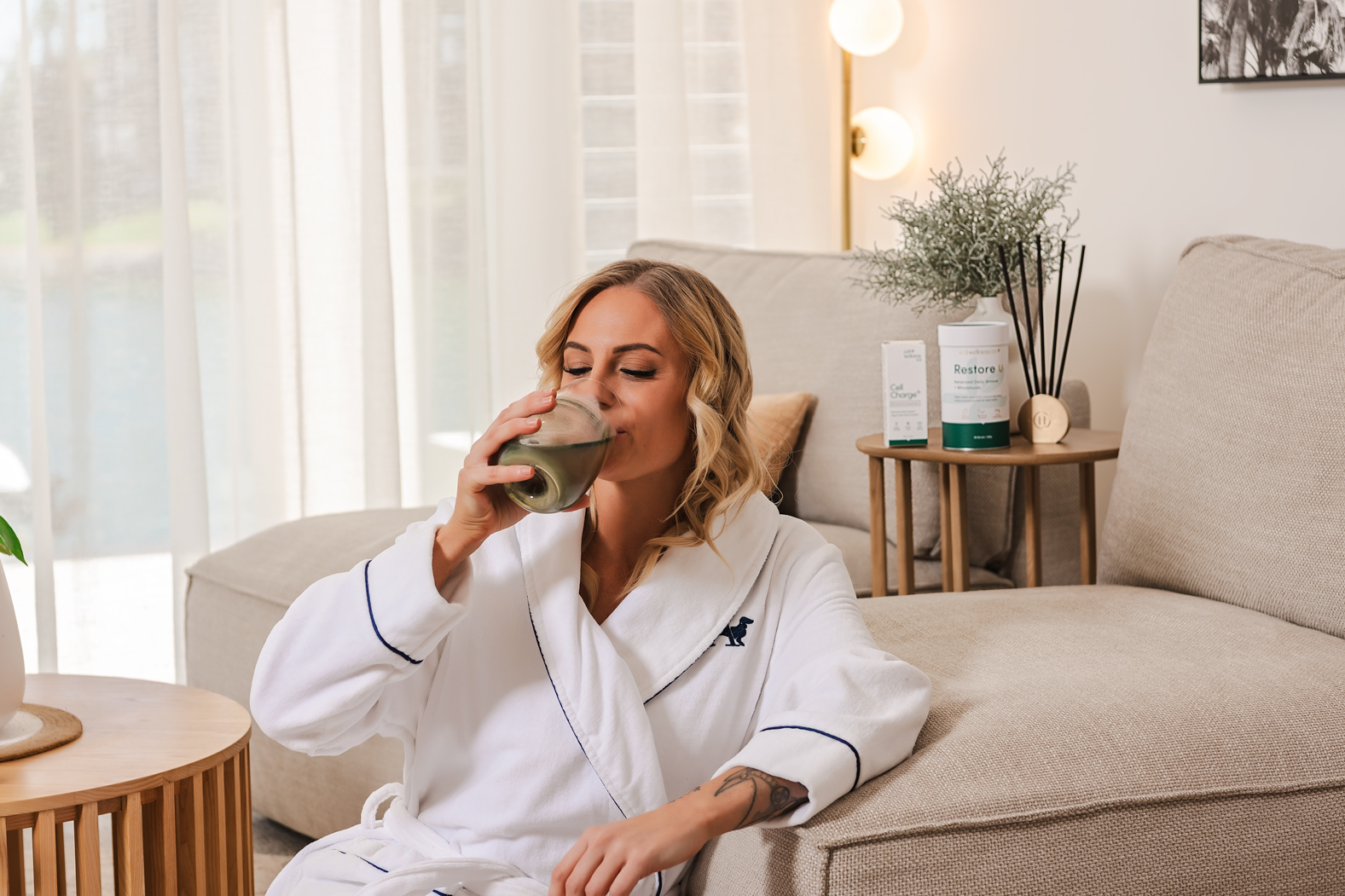Woman in a white robe drinking Restore U at home, with Restore U on the side table